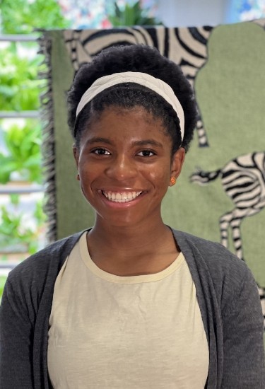 Zaila Avant-Garde smiles while wearing a white top with matching headband and a dark blue cardigan. Trees can be seen outside the window she is posing in front of